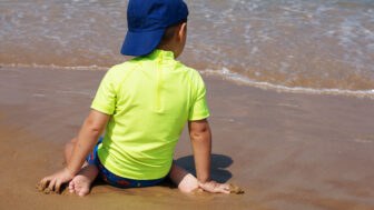 A small child in a yellow T shirt sits on his knees and looks at the sea